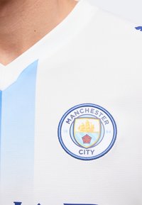 Εμβλημα του ποδοσφαιρικού συλλόγου Manchester City σε λευκή και γαλάζια ριγωτή φανέλα που φοράει ένα άτομο.