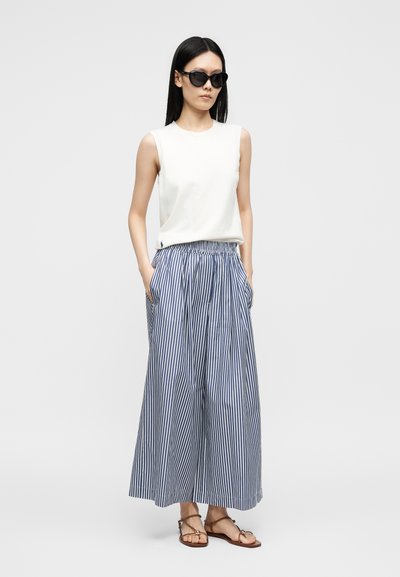 Femme portant des lunettes de soleil noires, un haut blanc sans manches, un pantalon large rayé bleu et blanc, et des sandales marron, debout, les mains dans les poches.