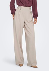 Μπεζ παντελόνι με ευρύ πόδι και πιέτες στη μέση, διακοσμημένο με ασημένιες κυκλικές μεταλλικές λεπτομέρειες. Το ύφασμα είναι απαλό και ελαφρώς ριγμένο.