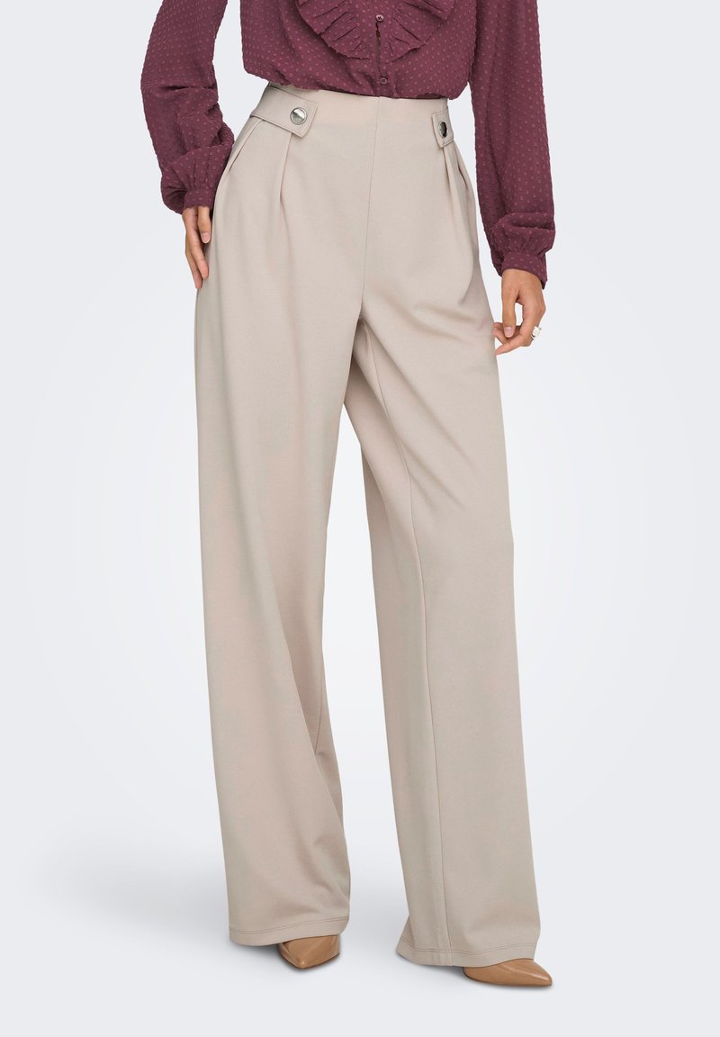 Μπεζ παντελόνι με ευρύ πόδι και πιέτες στη μέση, διακοσμημένο με ασημένιες κυκλικές μεταλλικές λεπτομέρειες. Το ύφασμα είναι απαλό και ελαφρώς ριγμένο.