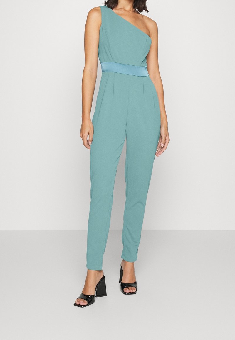 Femme portant une combinaison teal à une épaule avec une ceinture en satin, associée à des sandales à talons blocs noires ouvertes à l'avant, debout devant un fond blanc.
