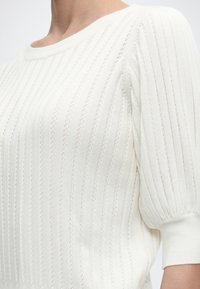 Nærbilde av en person iført en hvit ribbestrikket genser med korte puffede ermer og rund hals.