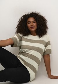 Tricou cu dungi în crem, cu dungi orizontale taupe, cu mâneci scurte și o croială lejeră, purtat cu pantaloni negri, stând sprijinit de un perete alb.
