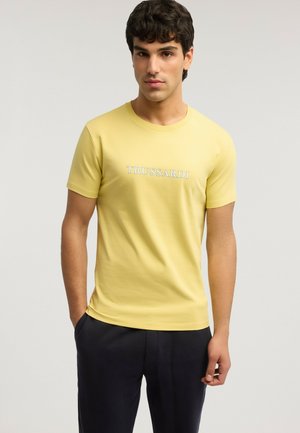 Junger Mann mit dunklem, lockigem Haar, der ein gelbes Trussardi-T-Shirt und schwarze Hosen trägt und mit einer Hand in der Tasche steht.