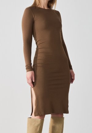 Kvinde iført en tætsiddende brun langærmet kjole med en sideslids, kombineret med beige knæhøje støvler mod en ensartet lys baggrund.