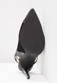 Chaussure à talon haut noire à bout pointu inclinée contre un mur blanc d’angle, montrant la semelle lisse et un petit talon stiletto.