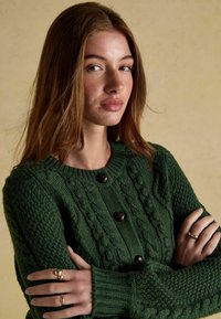 Zielony sweter robiony na drutach z teksturowanym wzorem w warkocze, z okrągłym dekoltem i brązowymi guzikami. Grube mankiety nadają konstrukcję projektowi.