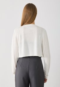 Cropped, lightweight white long-sleeve top with a round neckline, worn with high-waisted dark gray trousers. Smooth texture, minimal design.