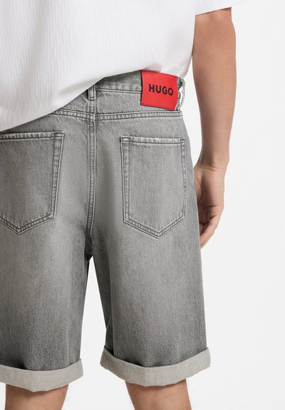 Shorts en denim gris avec revers roulés et poches arrière, arborant une étiquette rouge avec le nom de la marque "HUGO" sur la ceinture.