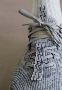 Nærbillede af en grå strikket sneakersko med lysegrå snørebånd bundet i en sløjfe og et mønstret trækstykke på en beige overflade.