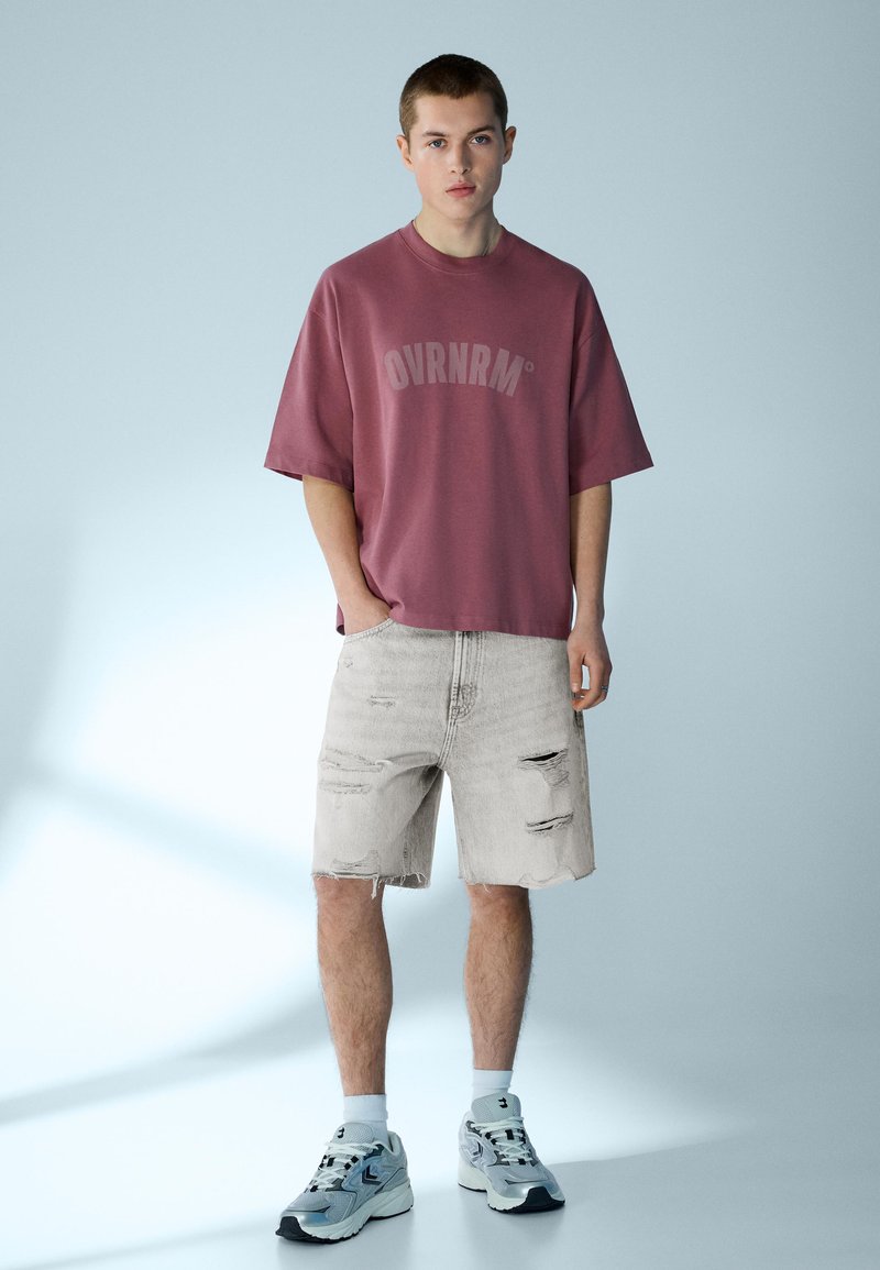 Jeune homme debout portant un t-shirt mauve ample avec le texte « OVRNRM », un short en jean clair déchiré, des chaussettes blanches et des baskets grises, sur un fond pâle.
