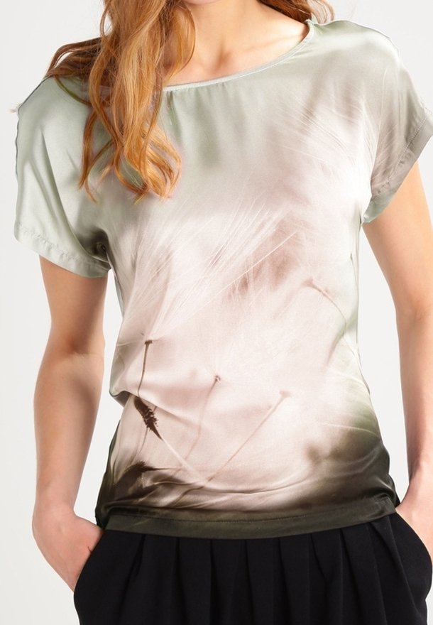 Woman wearing a soft green short-sleeve top with a delicate dandelion seed print and black pleated pants, hands in pockets.