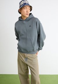 Grau Baumwollhoodie mit einer Kängurutasche und schwarzem Jordan-Logo. Kombiniert mit hellbraunen Hosen. Das Model trägt einen blauen karierten Bucket Hat.