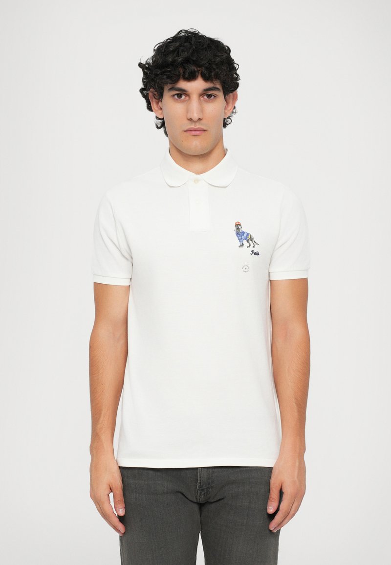 Camisa polo branca com colarinho, mangas curtas, apresentando um cão bordado com um suéter xadrez azul no lado esquerdo do peito.