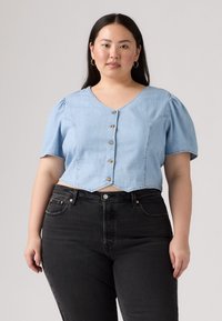 Camicetta di jeans cropped azzurra chiara con maniche corte a palloncino, scollatura a V e bottoni di legno sulla parte anteriore, abbinata a jeans neri scuri.