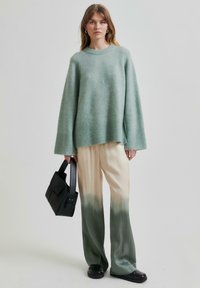 Mátově zelený oversized svetr s žebrovanými manžetami, spárovaný s krémovými a zelenými ombré širokými kalhotami. Drží černou kabelku.