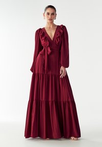 Bordo spalvota maksi suknelė su raukinėliu ant kaklo ir ilgomis rankovėmis, turinti rišamą juosmenį ir sluoksniuotą sijoną iš minkštos medžiagos.