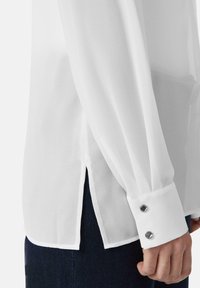 Witte doorschijnende blouse met lange mouwen, zijsplitten en knoopmanchetten met metalen accenten; gladde textuur en lichte stof.