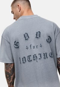 Szara, oversize'owa koszulka z miękkiego materiału, z ciemnym tekstem w dekoracyjnym kroju na plecach. Krótkie rękawy i luźny krój.