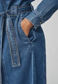 Modrá džínsová šaty s opaskom v páse, obsahujúce vrecko a detail s gombíkovým manžetou. Tkanina vykazuje klasickú textúru denimu.