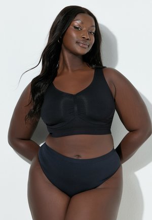 Selbstbewusste Frau mit langen Haaren, die einen schwarzen, ärmellosen Sport-BH und hoch taillierte Unterwäsche trägt und vor einer schlichten, hellen Wand steht.