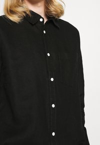 Camicia button-up nera in lino con colletto, dotata di bottoni bianchi e taschino sul petto. La texture è leggermente stropicciata, conferendo un aspetto rilassato.