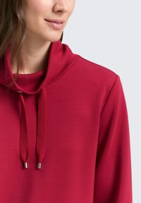 Rotes Sweatshirt mit Kapuze, verstellbaren Kordeln mit metallischen Enden und einer weichen, glatten Textur.