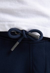 Marineblauwe broek met een gestructureerde tailleband, uitgerust met een donkerblauwe trekkoord die in een strik is geknoopt. Een wit shirt piept er bovenuit.
