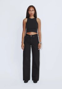 Frau mit langen Haaren, die ein schwarzes ärmelloses Croptop und eine schwarze, hoch taillierte, weite Hose trägt, steht vor einem schlichten, hellen Hintergrund.