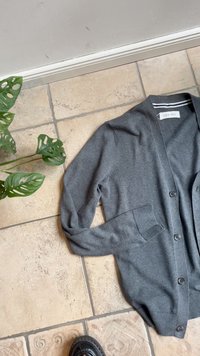 Grauer Cardigan aus weichem Stoff, mit V-Ausschnitt, Knopfverschluss und seitlichen Taschen, auf texturierten Fliesen ausgebreitet.