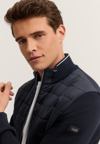 Młody mężczyzna z krótkimi brązowymi włosami nosi granatową pikowaną kurtkę z suwakiem i białą koszulkę pod spodem, pozując na jednolitym jasnym tle.