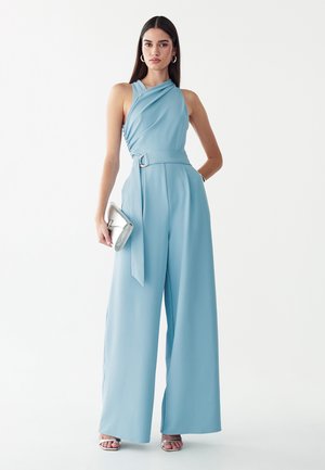 Frau trägt einen ärmellosen hellblauen Jumpsuit mit weitem Bein und Gürtel, hält eine silberne Clutch und trägt silberne offene High Heels.