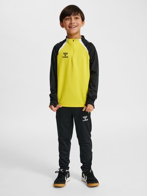 Smilende dreng, der står iført en gul og sort sportjakke og sorte træningsbukser med sorte sneakers på hvid baggrund.