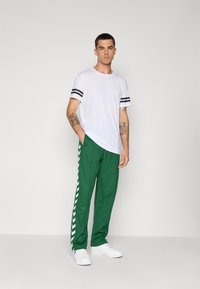 Witte t-shirt met zwarte strepen op de mouwen, gecombineerd met groene broek met een wit chevronpatroon langs de zijkanten. Witte sneakers maken de look compleet.