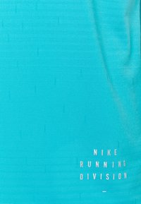 Tyrkysový běžecký top Nike, vyrobený z prodyšné texturované tkaniny, s bílým logem "NIKE RUNNING DIVISION" vytištěným blízko spodního lemu.