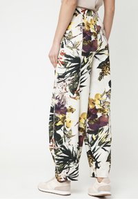 Vrouw in wijde bloemenprintbroek en beige sneakers, staand tegen een effen witte achtergrond.