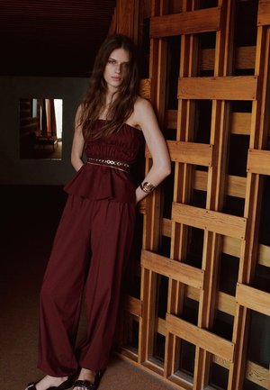 Donna con capelli lunghi e ondulati, che indossa una tuta smanicata color bordeaux e un braccialetto, appoggiata a una parete di graticcio in legno all'interno.