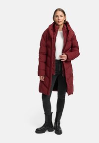 Bordeaux pufferjakke med hetta, glidelås og quiltet mønster. Bært over en hvit topp og svarte jeans, kombinert med svarte ankelboots.