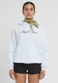 Sweat à capuche blanc avec une poche avant et le texte "Angel Face" en écriture noire. Agrémenté d'une écharpe à motifs verts nouée autour du cou.