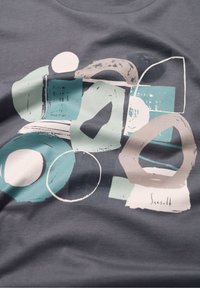 Pilkos medvilnės marškinėliai su abstrakčiu dizainu, kuriame yra apskritimų ir geometrinių formų derinys, sukurtas mėlynos, baltos ir smėlinės spalvų. Minkšta tekstūra su grafiniu spaudiniu.