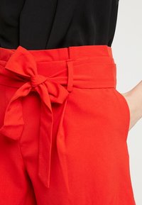 Czerwone spodnie z wysokim stanem i paskiem zawiązanym w kokardę. Gładka tkanina z bocznymi kieszeniami, dopasowany krój i szerokie nogawki.