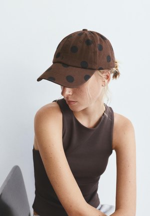 Žena u smeđoj majici bez rukava i smeđoj kapi s točkicama, gleda dolje i u stranu na jednobojnoj svijetloj pozadini.