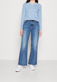 Modrá a bílá pruhovaná dlouhá košile s kulatým výstřihem, v kombinaci se světle modrými širokými džíny a bílými teniskami.