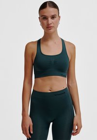 Donkergroene sportbeha met een geribbelde textuur, brede bandjes en een klein geraffineerd detail aan de voorkant, gecombineerd met bijpassende leggings.