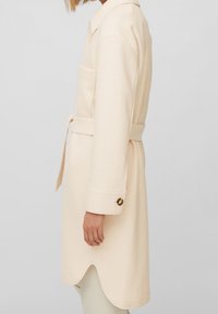 Abrigo de lana beige claro con cuello, bolsillos laterales y cinturón para ajustar a la cintura. Cuenta con un dobladillo festoneado y detalles de botones dorados en las mangas.