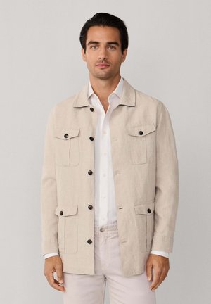 Man draagt een beige jasje met knopen en vier zakken aan de voorkant, en een wit overhemd, staand tegen een lichtgrijze achtergrond.