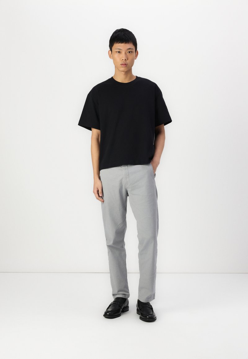 Camiseta de algodón negra de mangas cortas, combinada con pantalones gris claro y zapatos negros, sobre un fondo blanco.