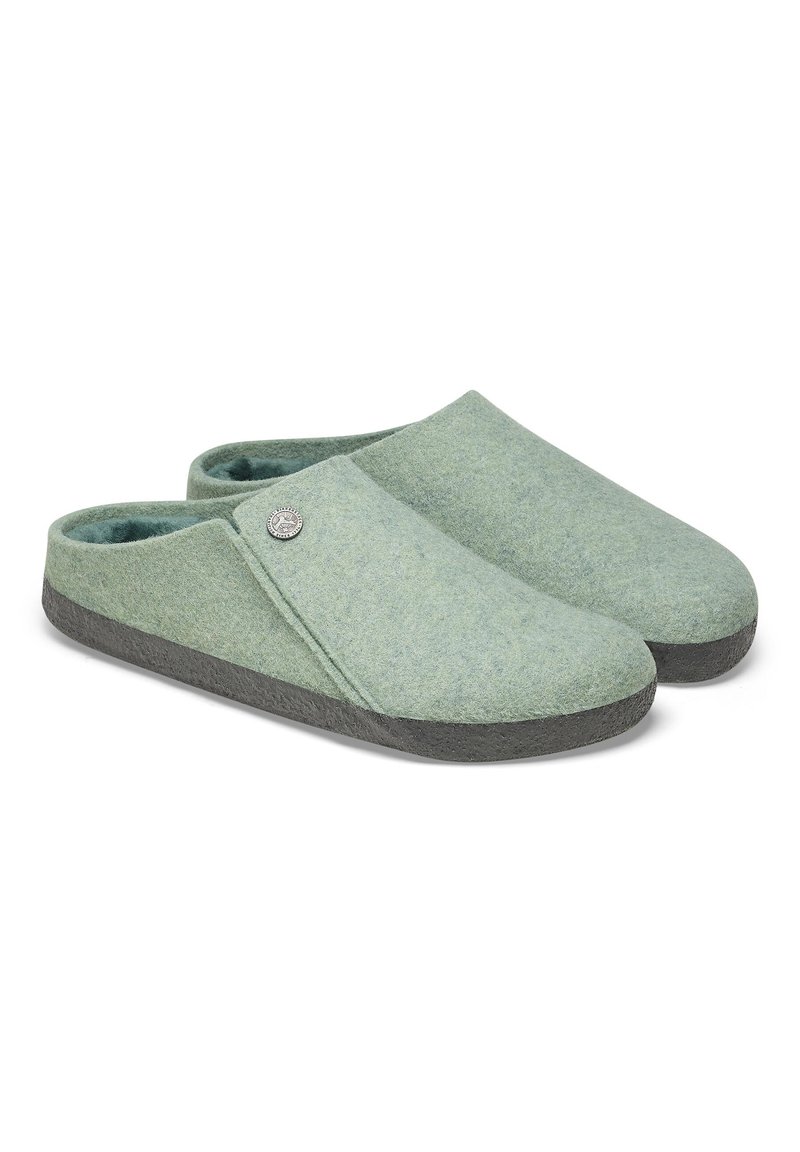 Grüne Filzschuhe mit abgerundetem Zehenbereich, gepolstertem Innenraum und dunkelgrauem Gummisohle. Merkmale eines runden Metallakzents auf dem Obermaterial.