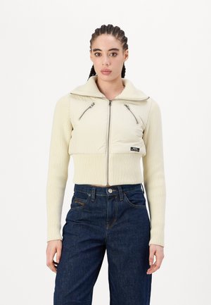 Diese taillierte Jacke hat ein cremfarbenes, gepolstertes Oberteil mit gerippten Ärmel, Reißverschlusstaschen und einem breiten Kragen. Sie wird mit dunklen Jeans kombiniert.