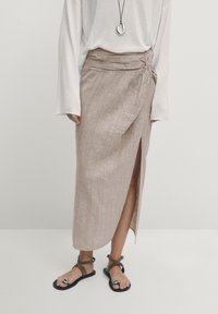 Jupe en lin marron clair avec une fente latérale et un détail froncé à la taille, portée avec des sandales plates noires et un haut blanc à manches longues.